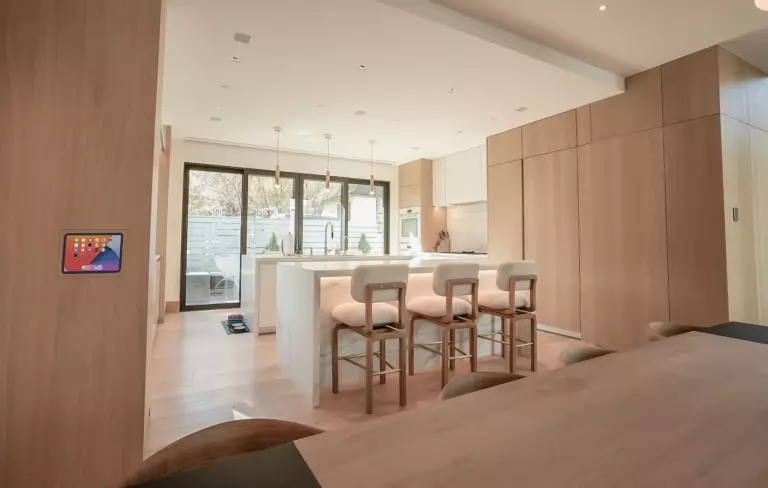 Modern kitchen with light wood cabinets, a white island with four barstools, pendant lights, and large windows overlooking a patio. A digital control panel is mounted on the wall.