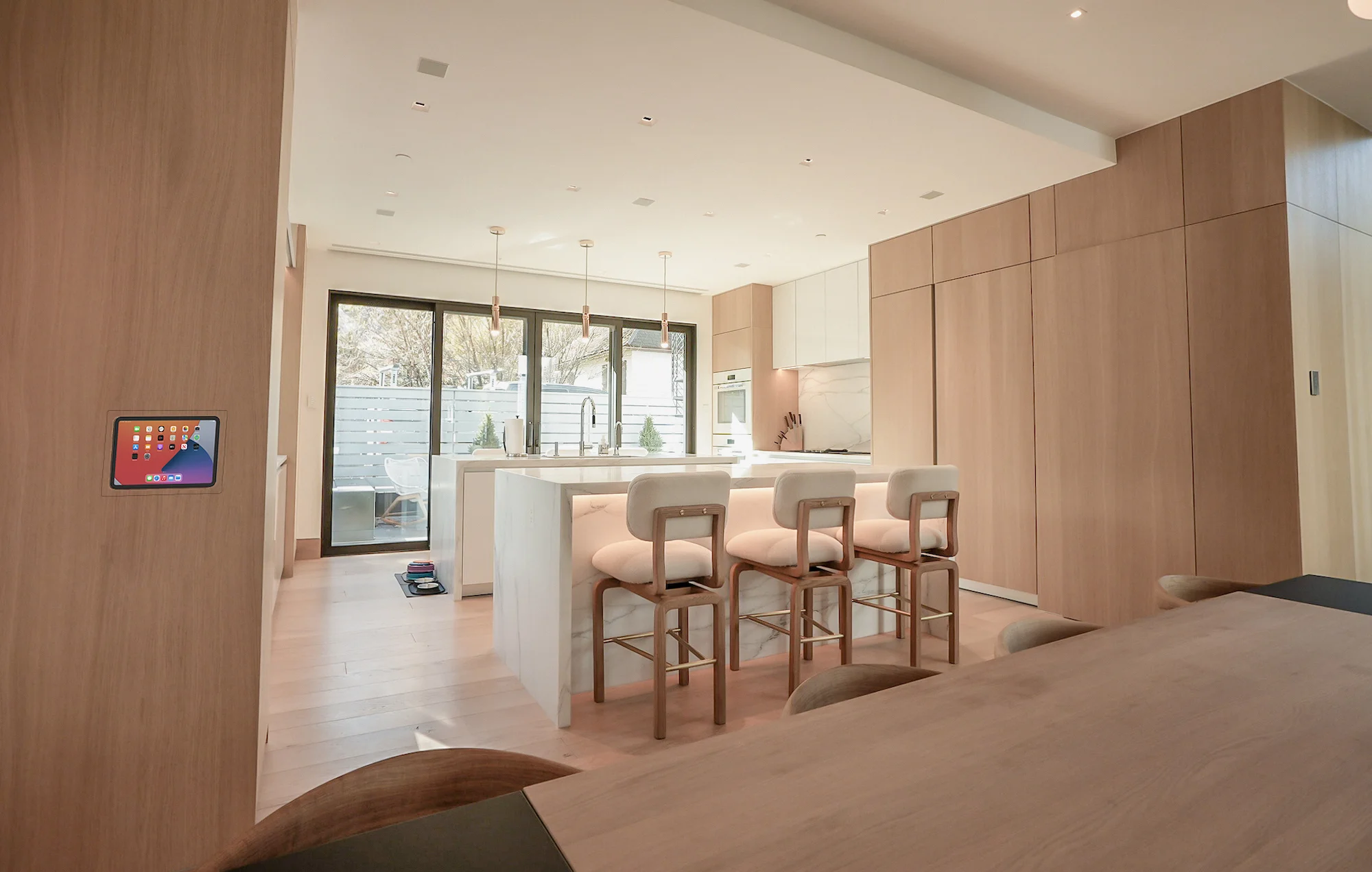 Modern kitchen with light wood cabinets, a white island with four barstools, pendant lights, and large windows overlooking a patio. A digital control panel is mounted on the wall.