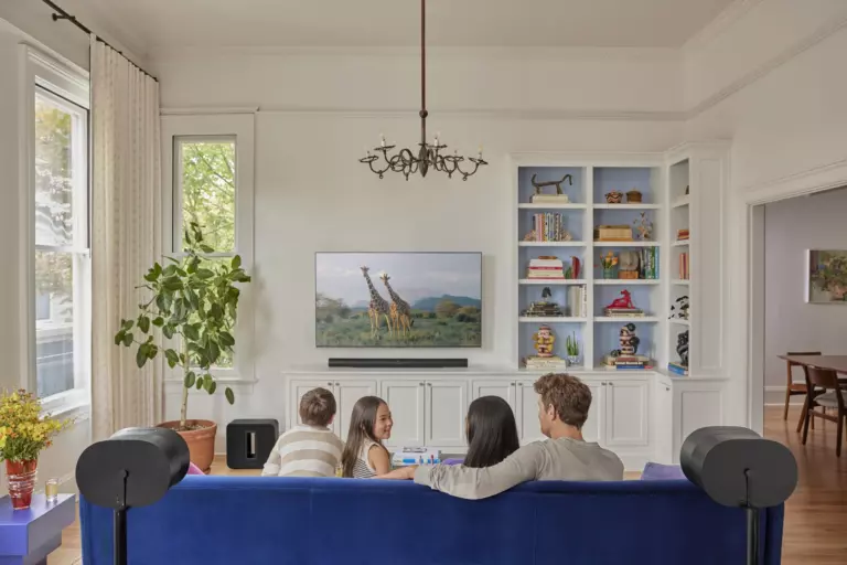 A family of four sits on a blue couch watching a nature show with giraffes on TV in a bright living room with bookshelves and plants.