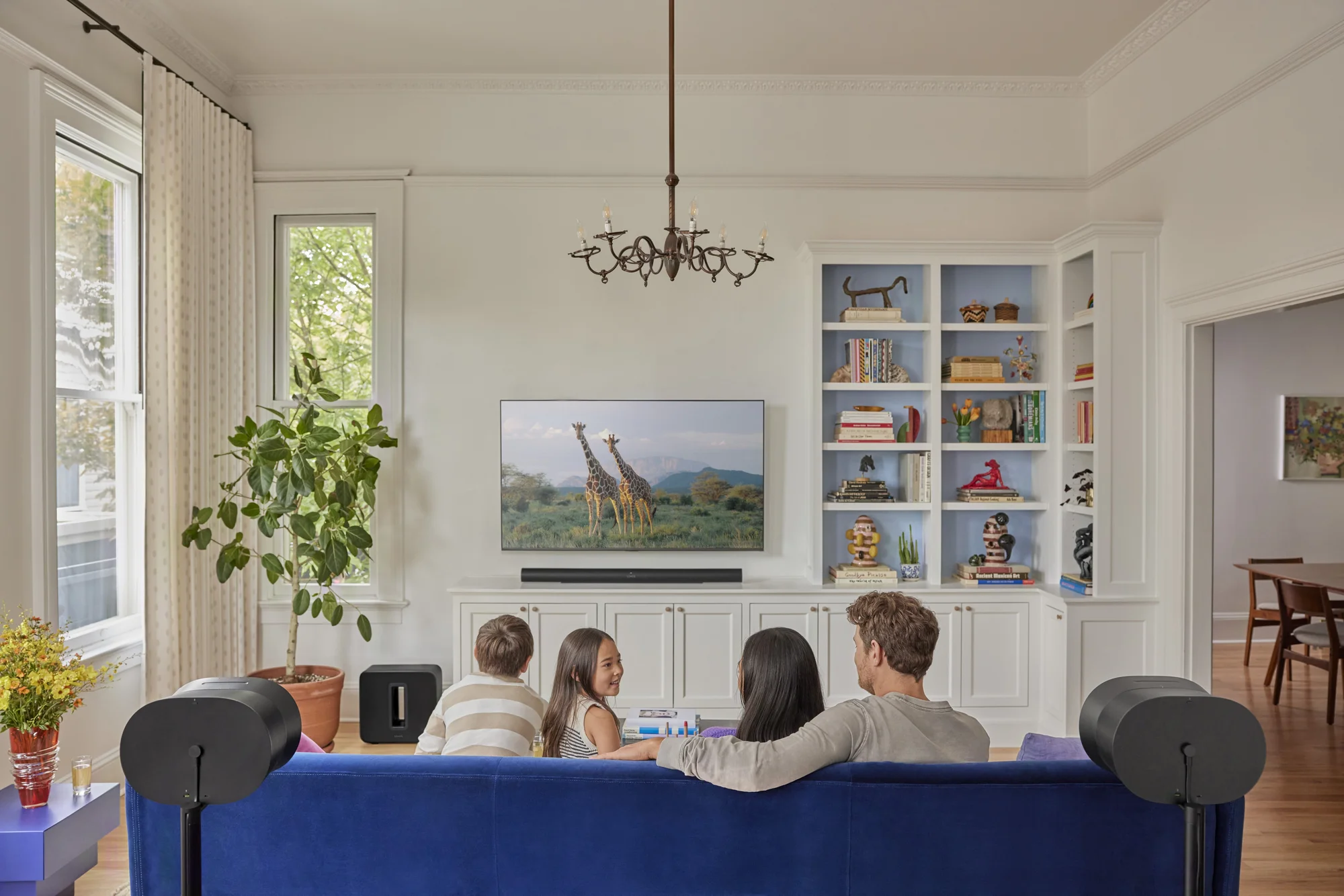 A family of four sits on a blue couch watching a nature show with giraffes on TV in a bright living room with bookshelves and plants.