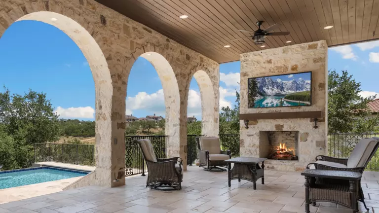 Covered stone patio with arched openings, seating, fireplace, mounted outdoor TV, and view of pool and landscape on a sunny day.