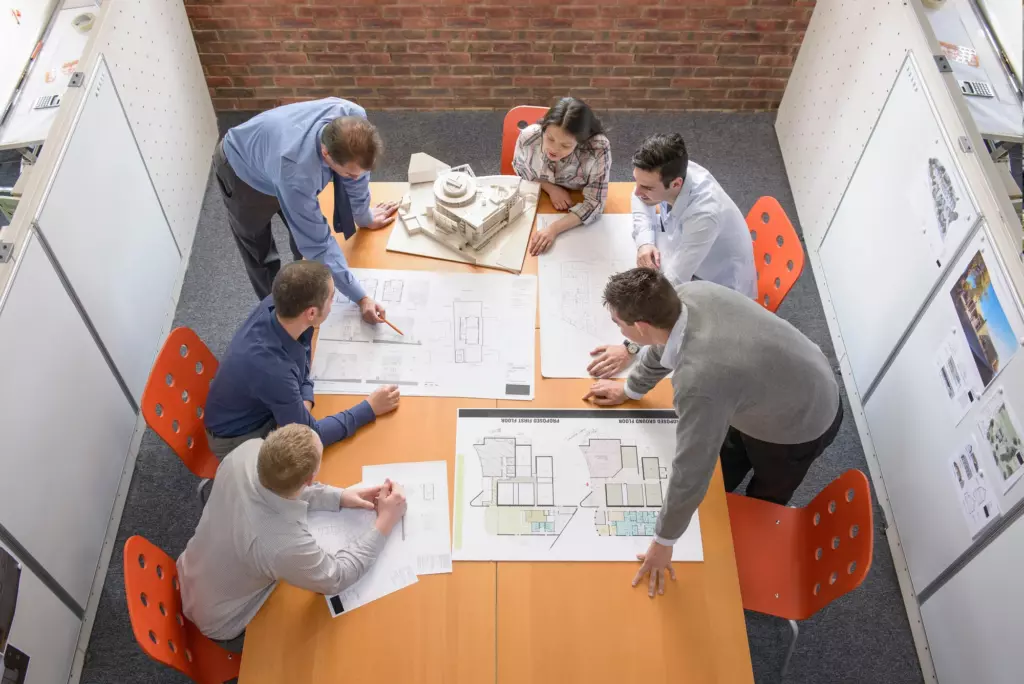 Six people gather around a table reviewing architectural blueprints and a building model in a modern office with whiteboards and brick walls.