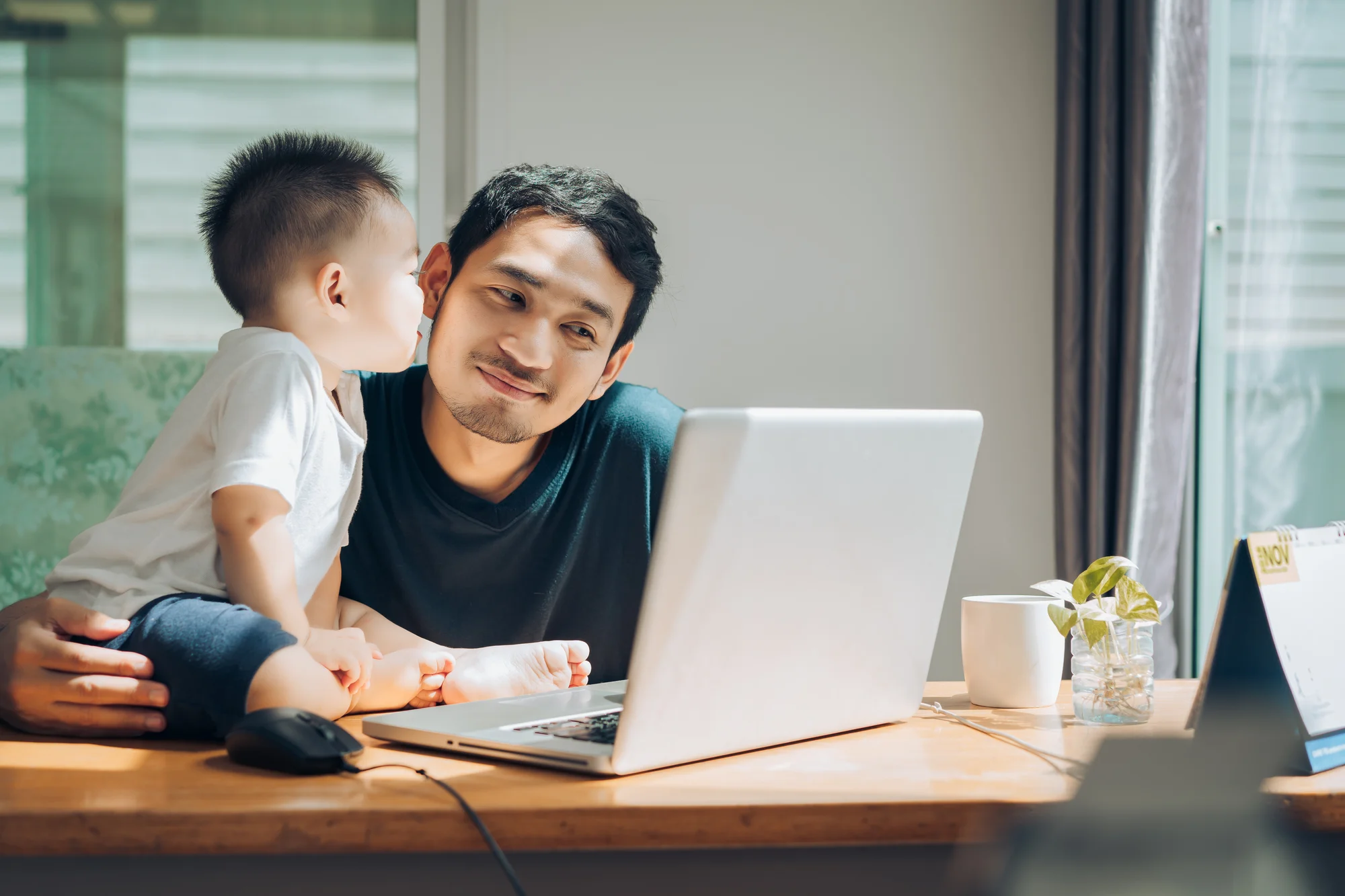 A man sits at a desk with a laptop, holding a child who is sitting on the desk and giving him a kiss on the cheek. Light streams in from a nearby window.