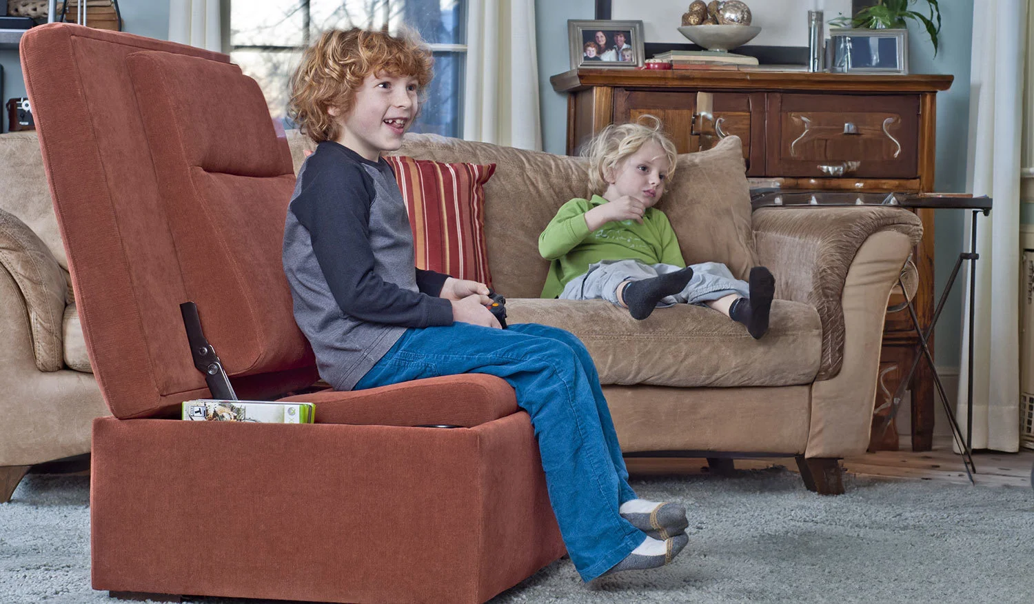 Two children watch TV in a living room; one sits on a red chair holding a video game controller, and the other lounges on a beige couch.