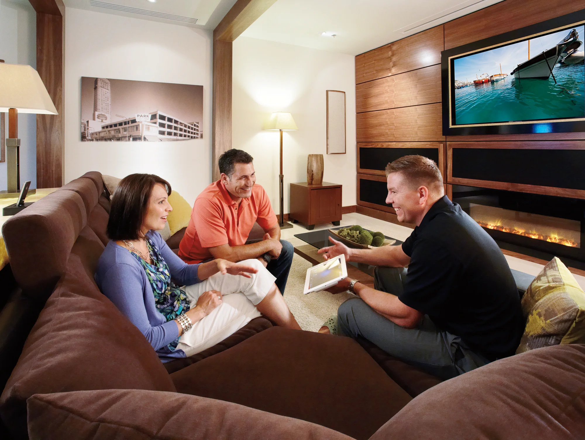 Three people sit on a brown sectional sofa in a modern living room, looking at a tablet while a boat is shown on the wall-mounted TV in the background.