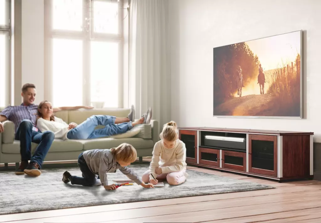 Two children draw on a rug while two adults relax on a couch in a bright living room with a large TV displaying a sunset scene.